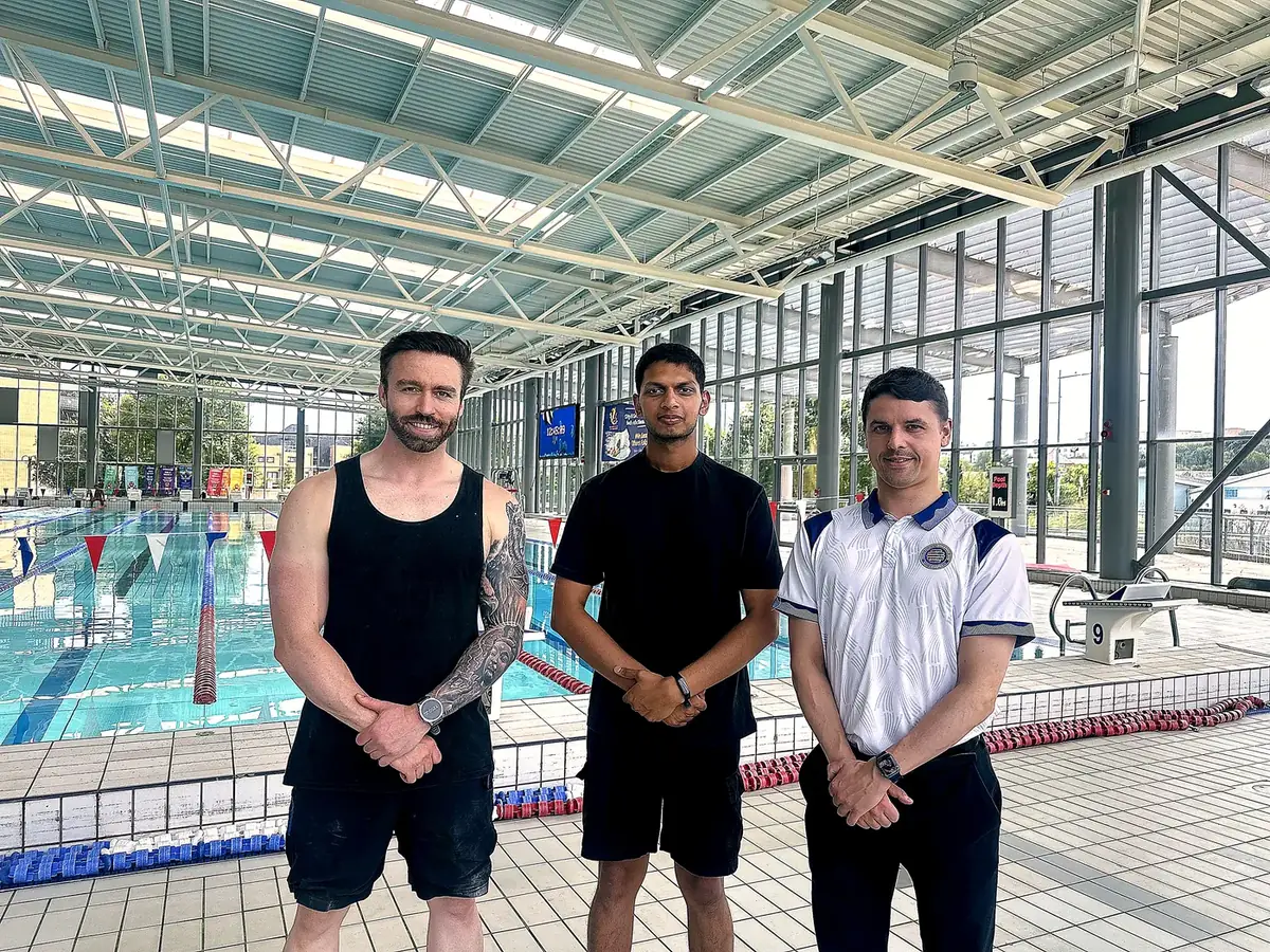 Three men standing in front of a fifty-metre indoor swimming pool